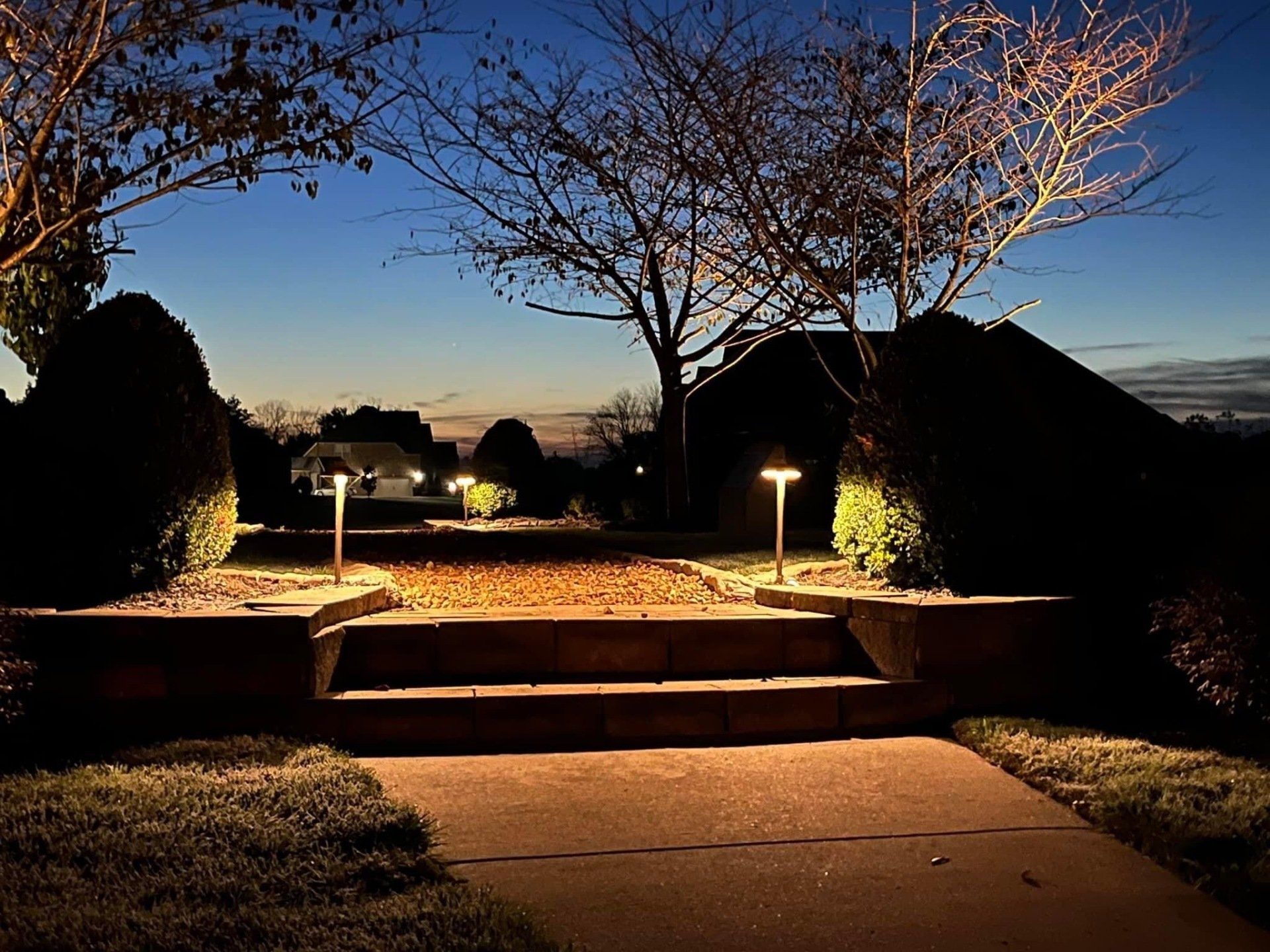 Evening walkway with steps, lit lampposts, trees, and dark shrubs under a twilight sky