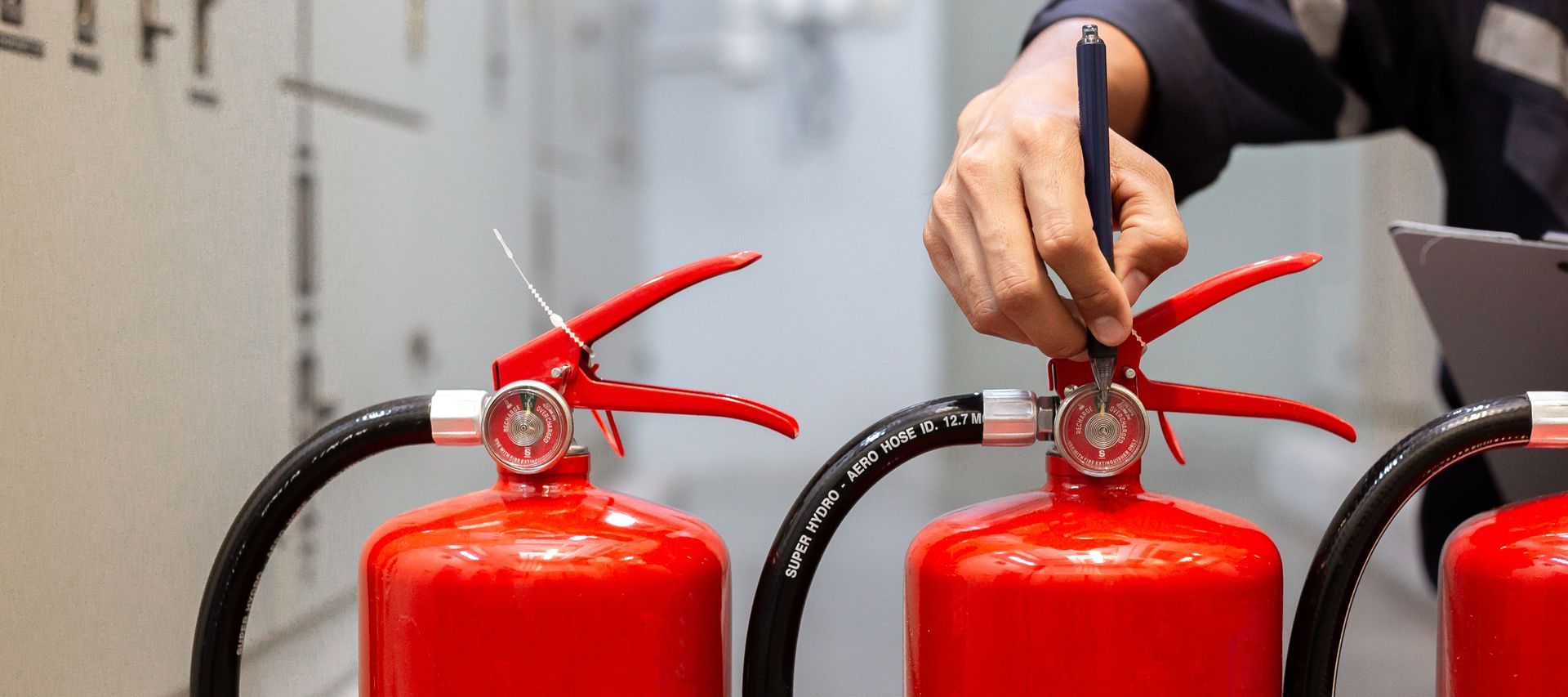 Technician's hand checking the pressure gauge on a row of red fire extinguishers inside a building.