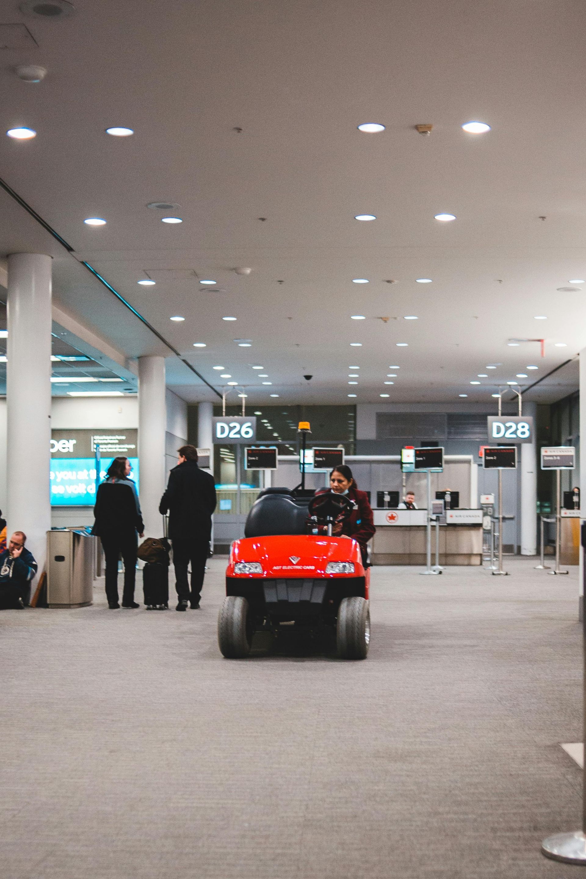 Un veicolo rosso con pneumatici sovradimensionati in transito in un aeroporto. La gente passa davanti ai banchi del check-in.