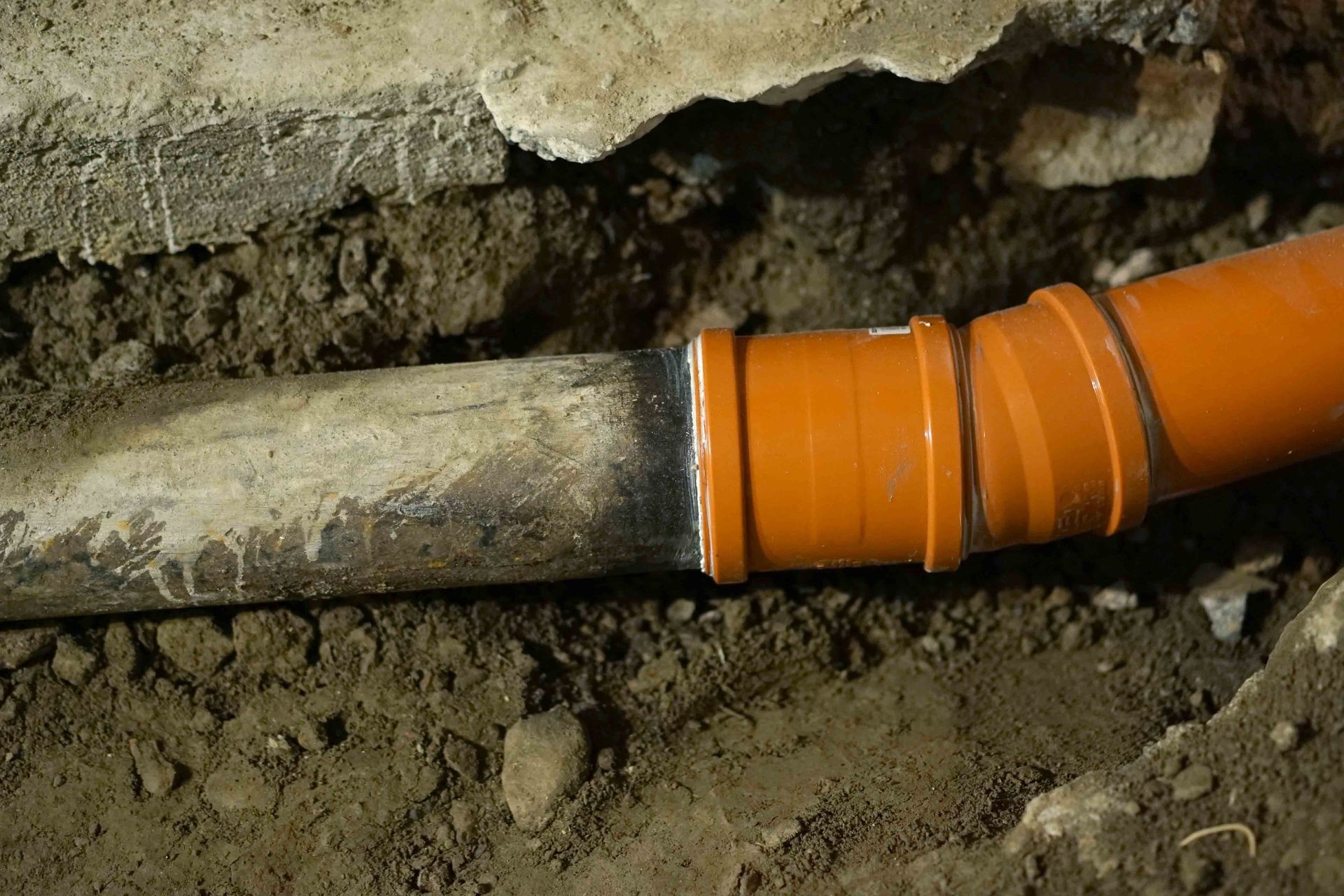 Ancien tuyau raccordé à un tuyau orange en PVC à l’aide d’un raccord, dans une excavation sous une dalle de béton.