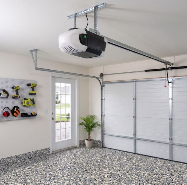Bright garage interior with a ceiling-mounted opener, closed white door, and wall-mounted tools near a side door.