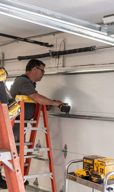 Technician on ladder installing garage door opener rail inside a garage, with tools and equipment nearby