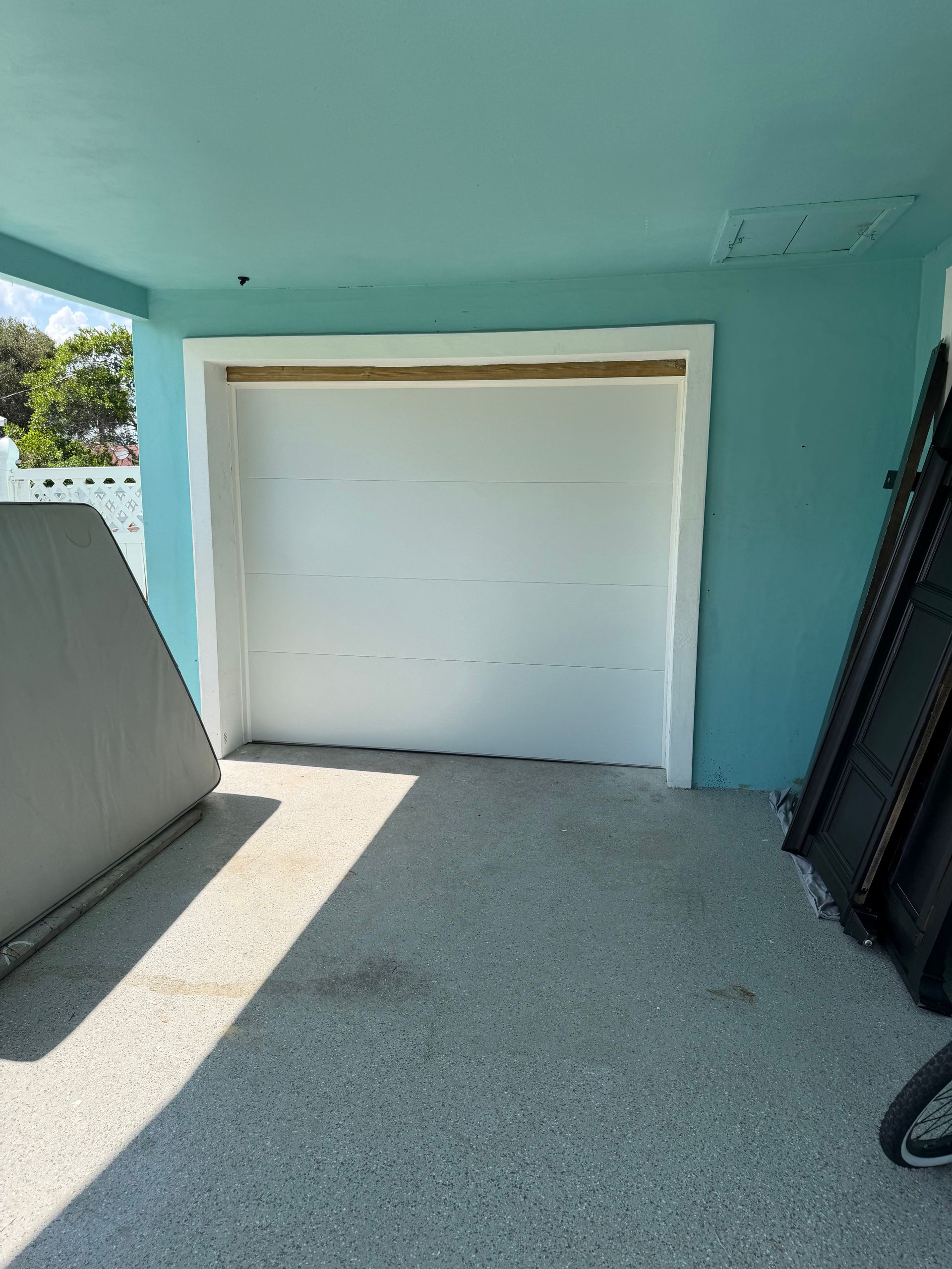 White garage door in a turquoise carport, with stacked items on both sides and sunlight on the concrete floor
