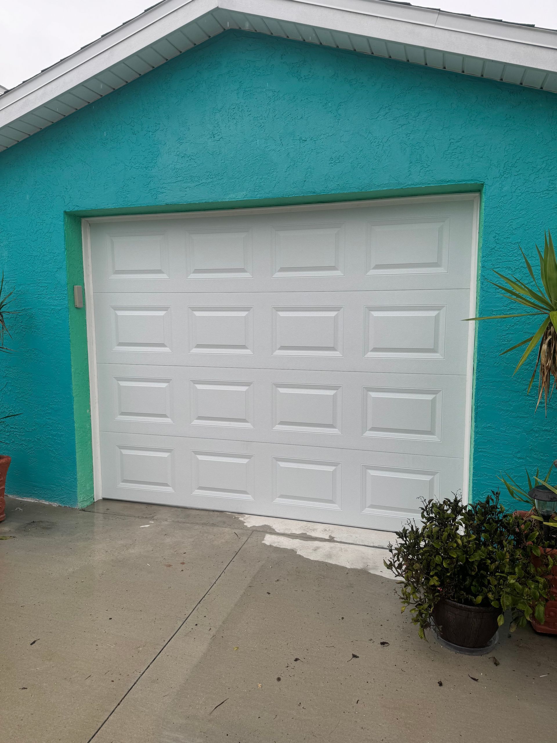 White garage door on a turquoise house with potted plants on a concrete driveway.