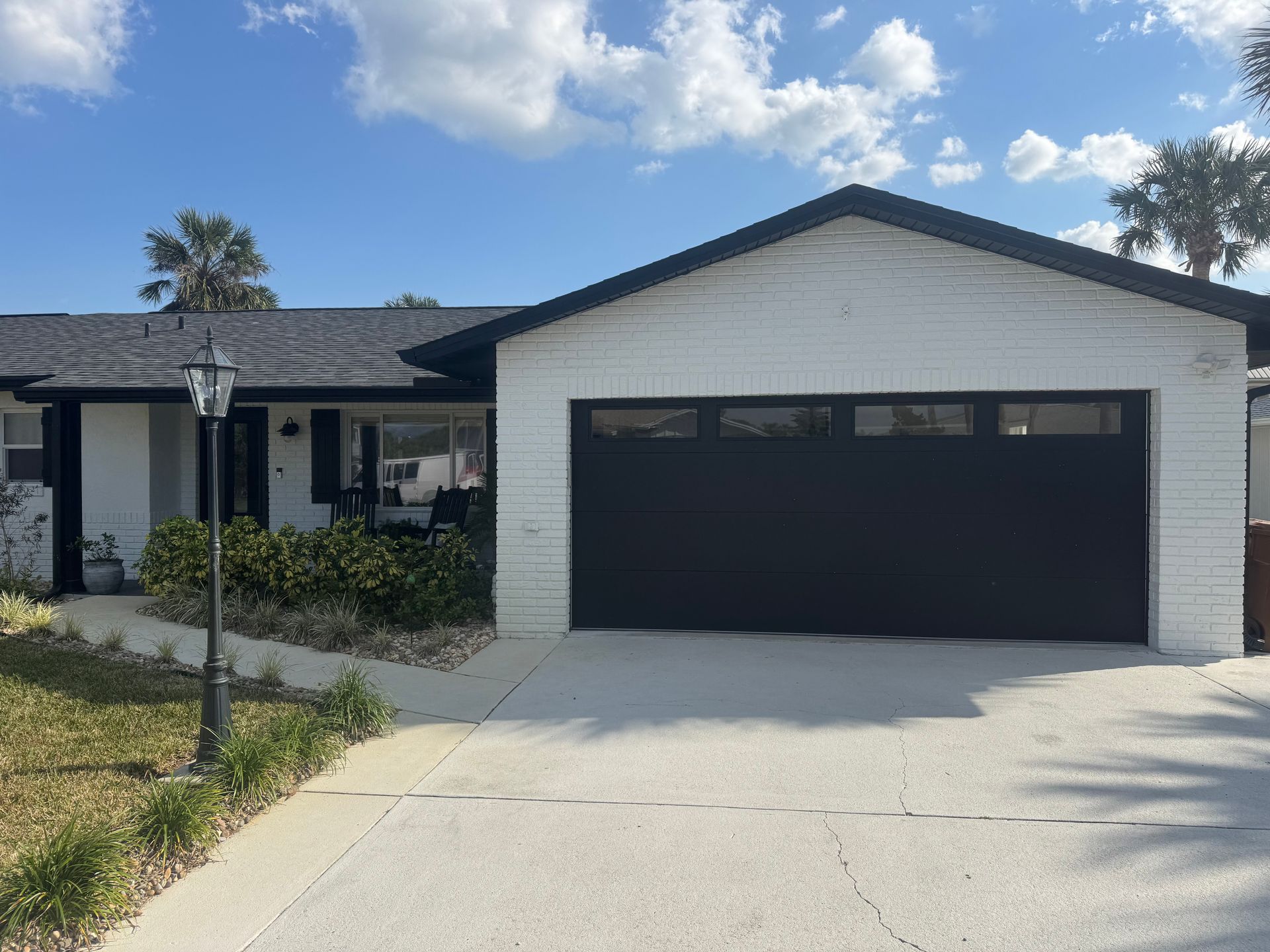 Single-story house with white brick, black garage door, and driveway under a blue sky.
