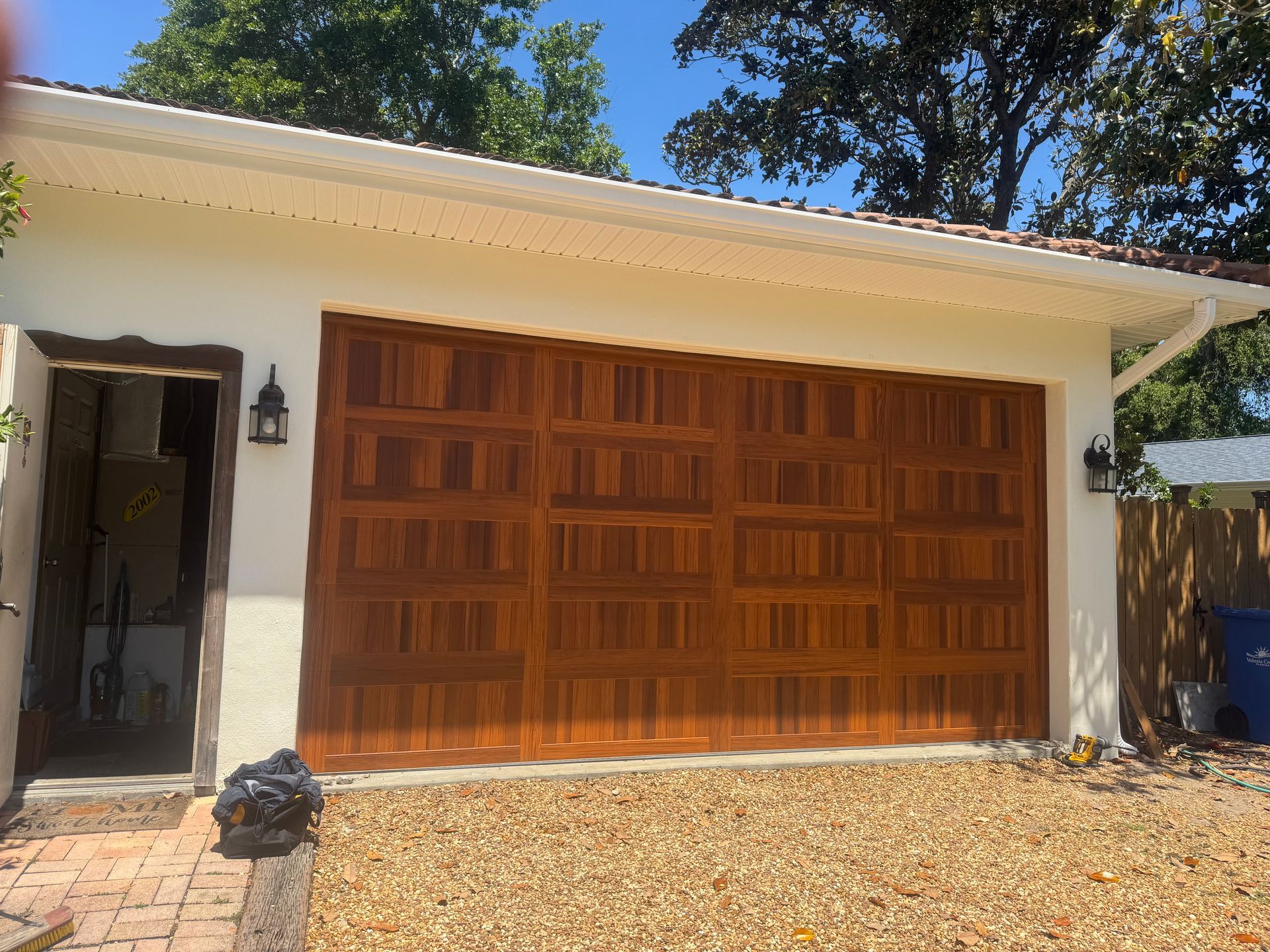 Single-story house with a wooden garage door, white trim, and a gravel driveway in bright sunlight
