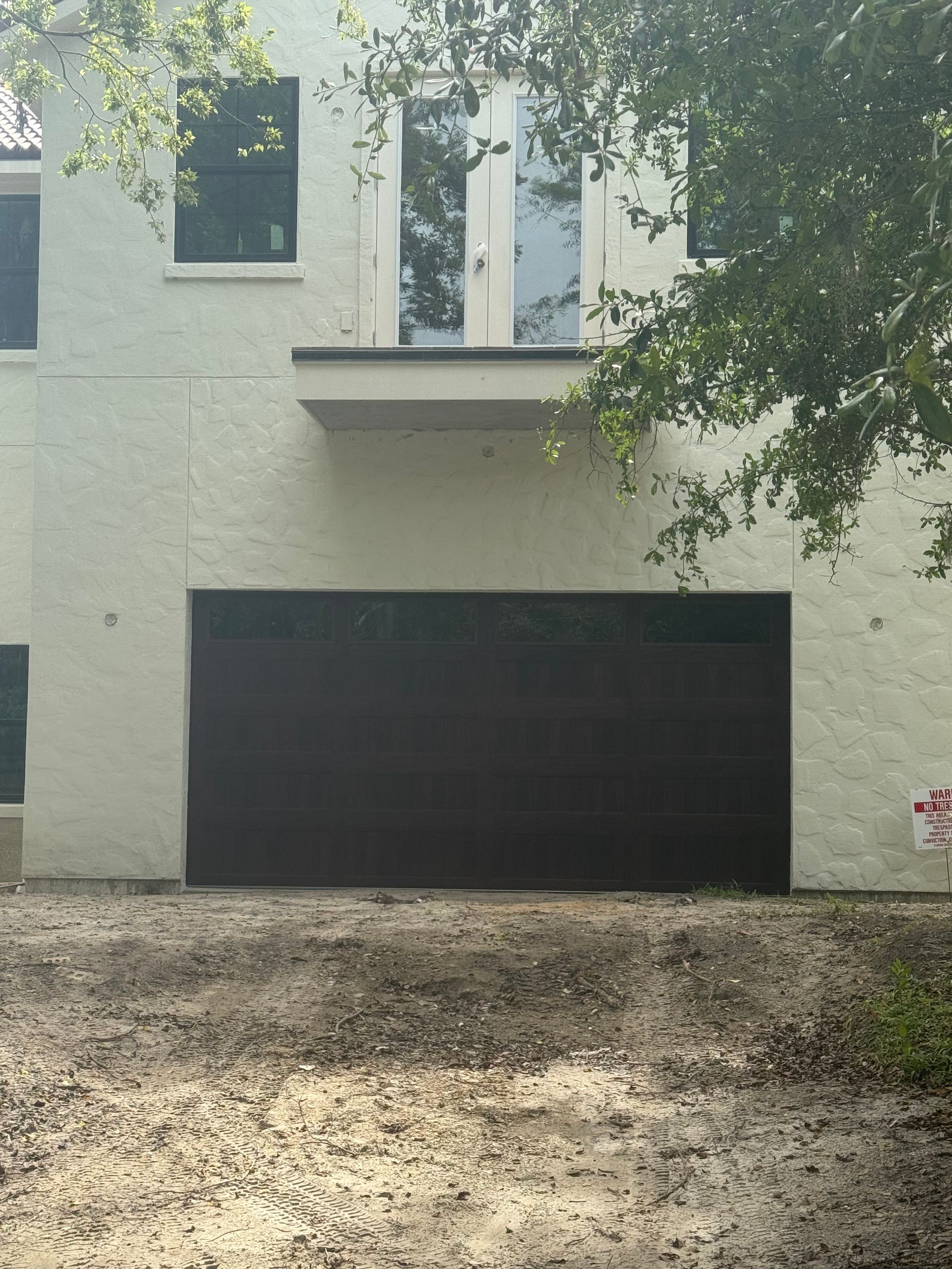 White house facade with black garage door, upper windows, and tree branches overhead