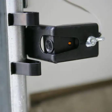 Black door hinge with a small security camera attached, mounted on a white wall.