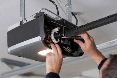 Technician adjusting a garage door opener with exposed wiring, viewed from below.