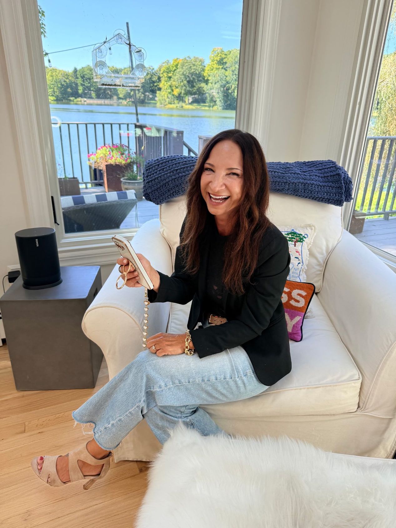 Amy woman seated on a white chair in a bright living room, holding a phone with a lake view behind her