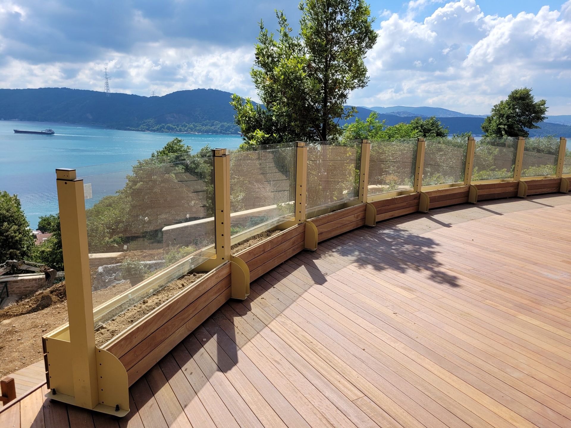 Curved wooden deck with glass railing overlooking a blue bay and green mountains under a partly cloudy sky