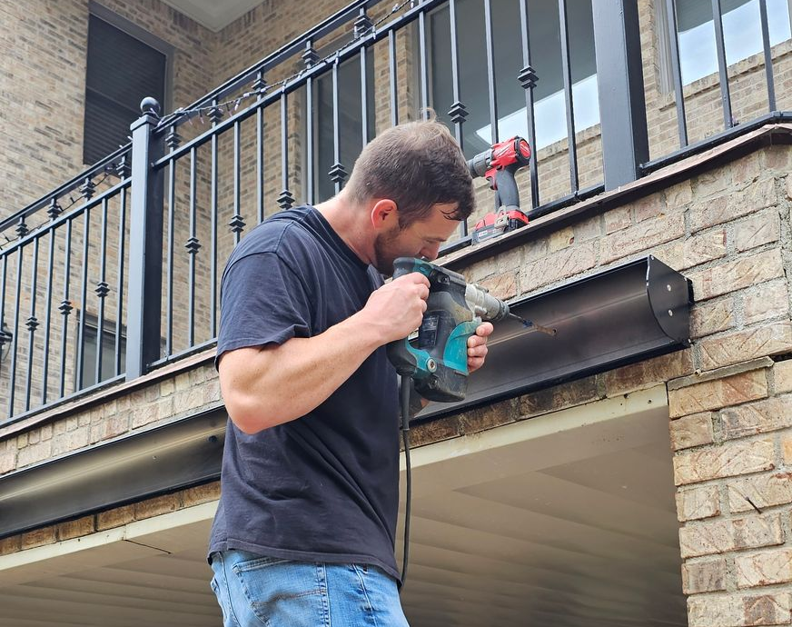 Man using a teal power drill to work on a metal gutter beneath a balcony