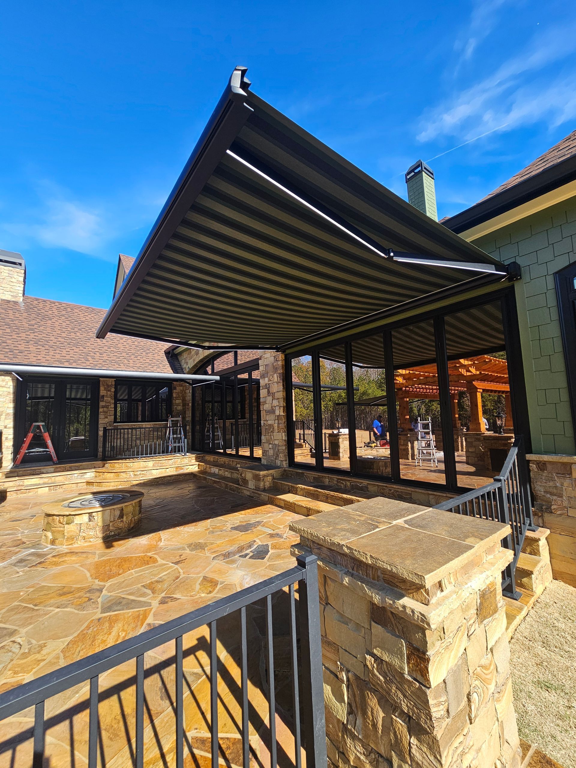 Covered stone patio with black metal railing and pergola beside a house under a blue sky