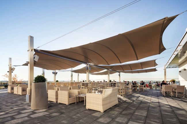 Outdoor café seating under large tan shade sails on a sunny terrace by the water