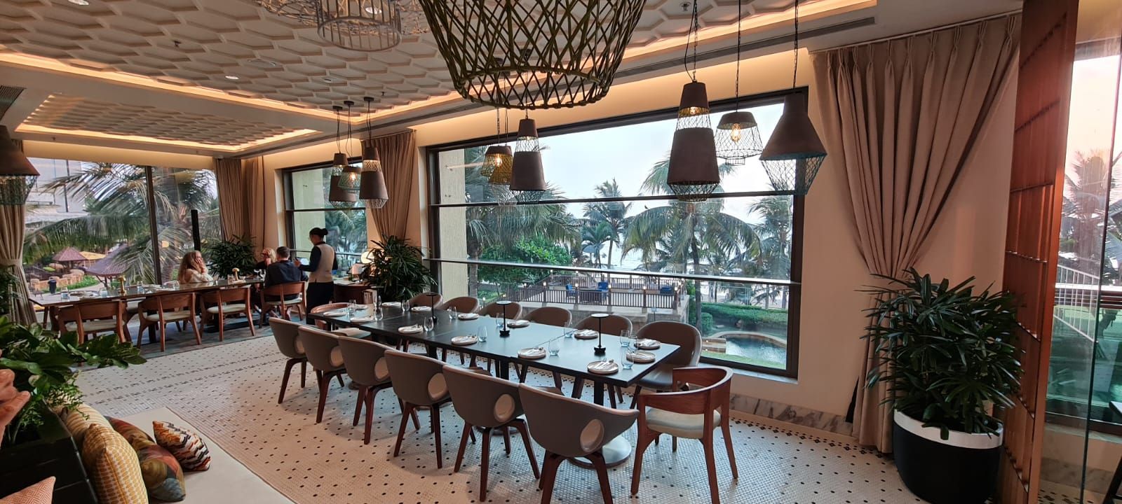 Elegant restaurant dining area with wooden tables, wicker chairs, large windows, and tropical greenery