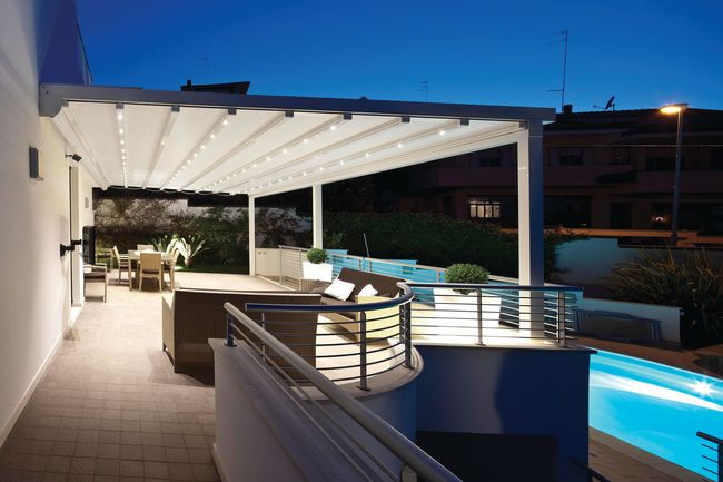 Modern patio at dusk with white pergola, curving pool, and city lights beyond