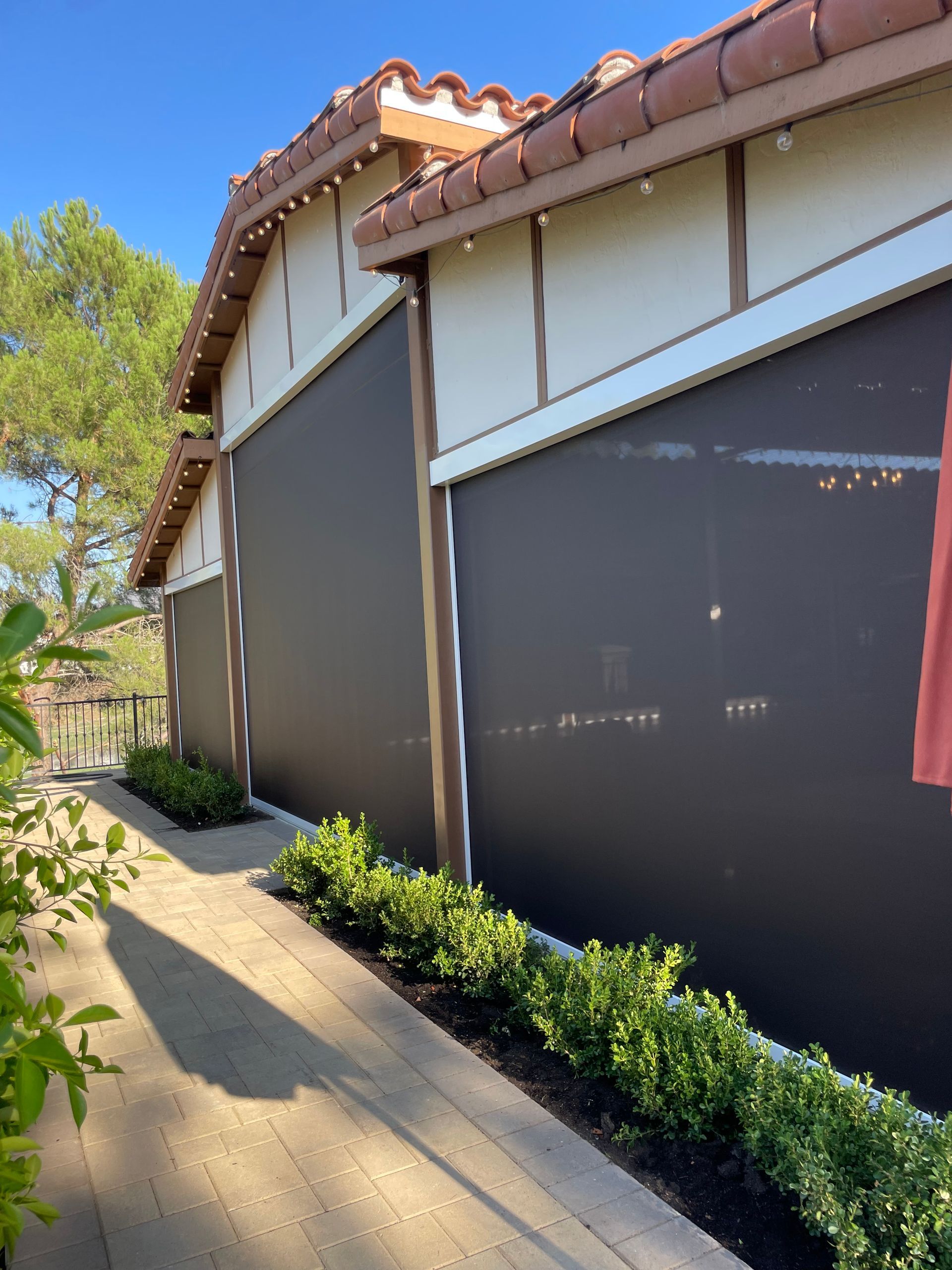 Outdoor walkway beside a stucco building with dark window shades, tiled roof, and trimmed shrubs under a blue sky