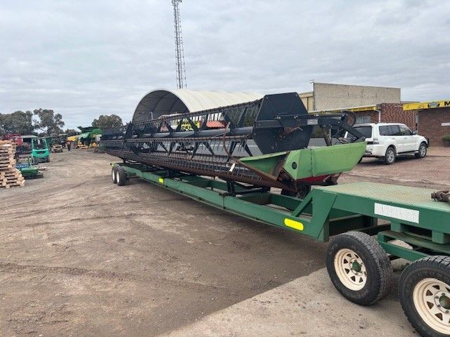 Green trailer carrying a large industrial machine in a yard, with a white pickup and warehouse in the background