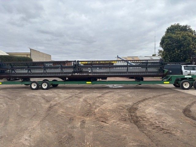 Long green agricultural trailer loaded on a semi-truck in a dirt lot under cloudy skies