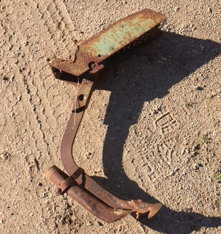 Rusty metal tool with a curved handle and blade, lying on sandy ground in sunlight