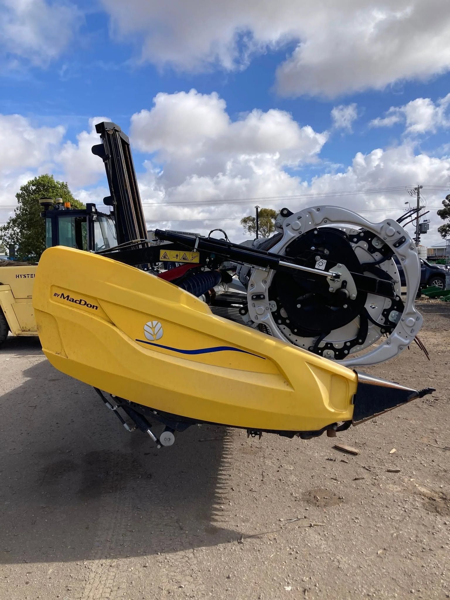 Yellow agricultural machine with large black wheel parked on gravel under a cloudy blue sky