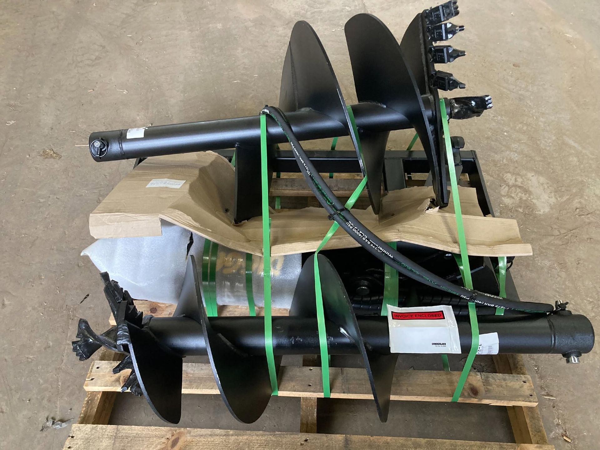 Two black auger drill attachments strapped to a cardboard pallet with green bands.
