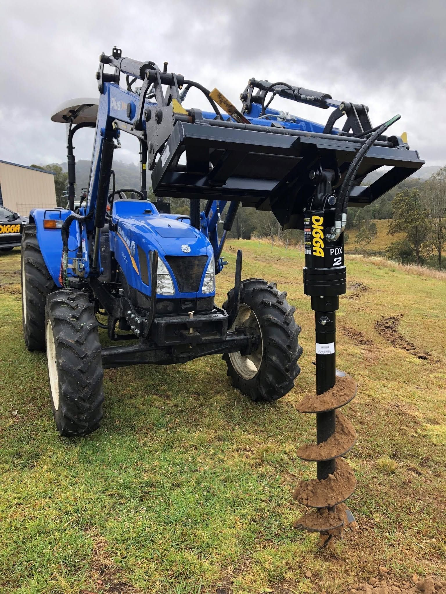 Blue tractor with auger attachment drilling into soil in a grassy field