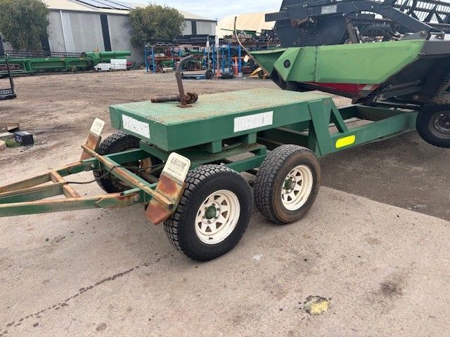 Green utility trailer with dual wheels parked beside a larger green machine in a lot