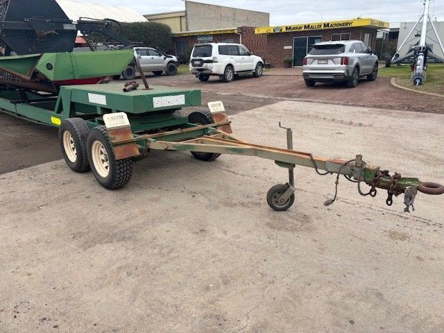 Green agricultural trailer with hitch and jack stand parked in a lot near trucks and buildings