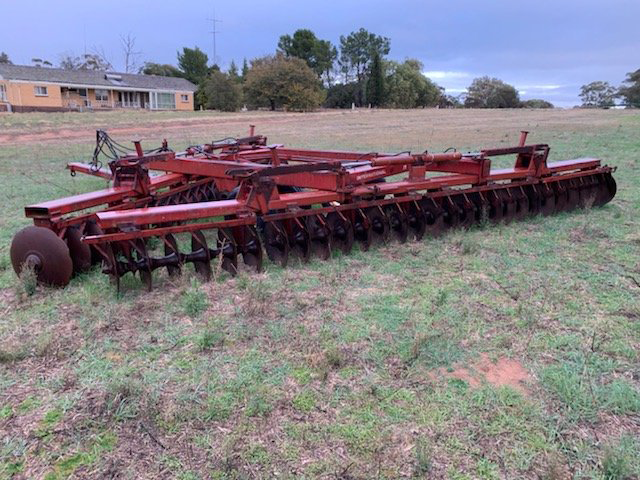 Red farm disc harrow with black blades parked in a grassy field near a house at dusk