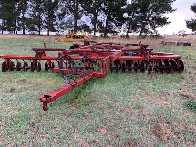 Red agricultural disc harrow parked on grass in a field, with trees in the background