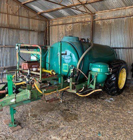 Green agricultural sprayer trailer inside a corrugated metal shed, with hoses and yellow wheels.