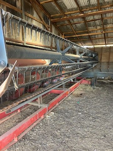 Interior of a barn with a long metal grain conveyor and red supports on a dirt floor