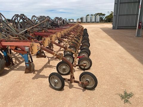Rows of farm tillage implements with wheels lined up on a gravel yard beside a metal shed