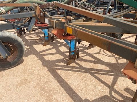 Tractor-towed farm implement with metal frame and blue support jacks on dirt ground in sunlight