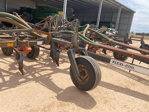 Farm machinery with large hoses and wheels parked on sandy ground outside a shed
