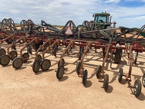 Rows of rusty farm implements with wheels in a dusty field, with a tractor in the background