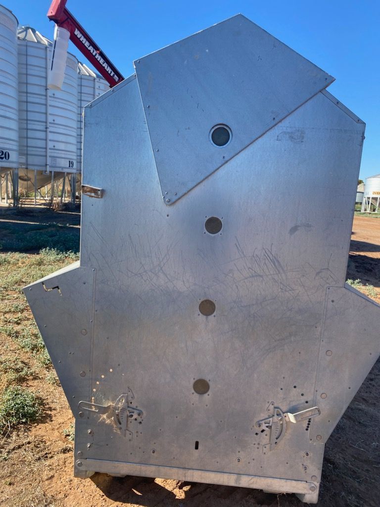 Silver metal industrial hopper with circular holes, outdoors on grass near a building.