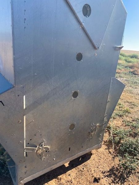 Close-up of a gray metal trailer or box with circular holes and a latch outdoors on dirt and grass