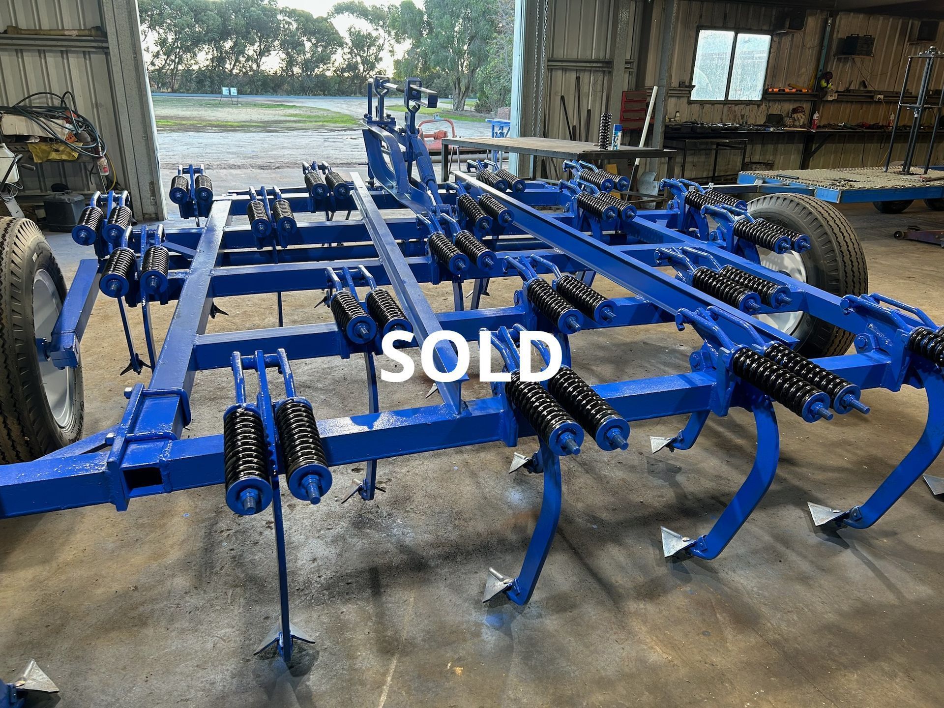 Blue farm implement with tines and wheels inside a shed, marked SOLD
