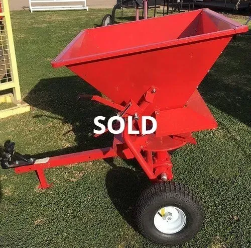Red agricultural hopper on a wheeled cart in a grassy yard, marked “SOLD”