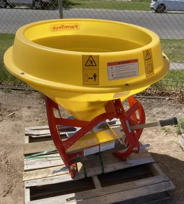Yellow agricultural spreader hopper on a red frame sitting on wooden pallets outdoors.