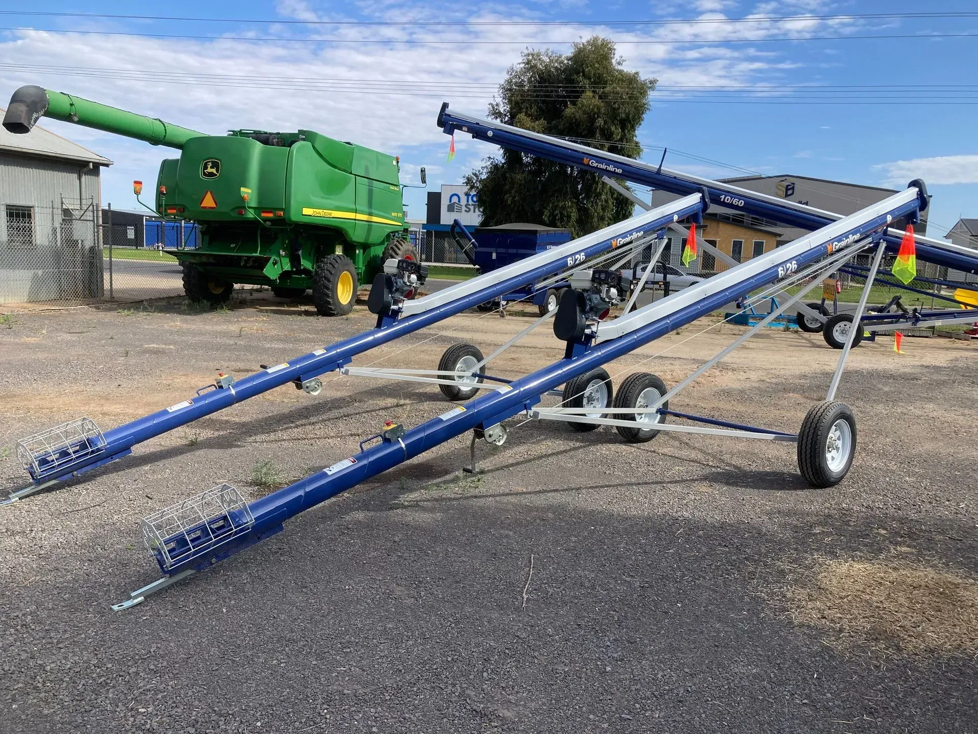 Blue agricultural auger on wheels in a farmyard, with green machinery and a person in the background