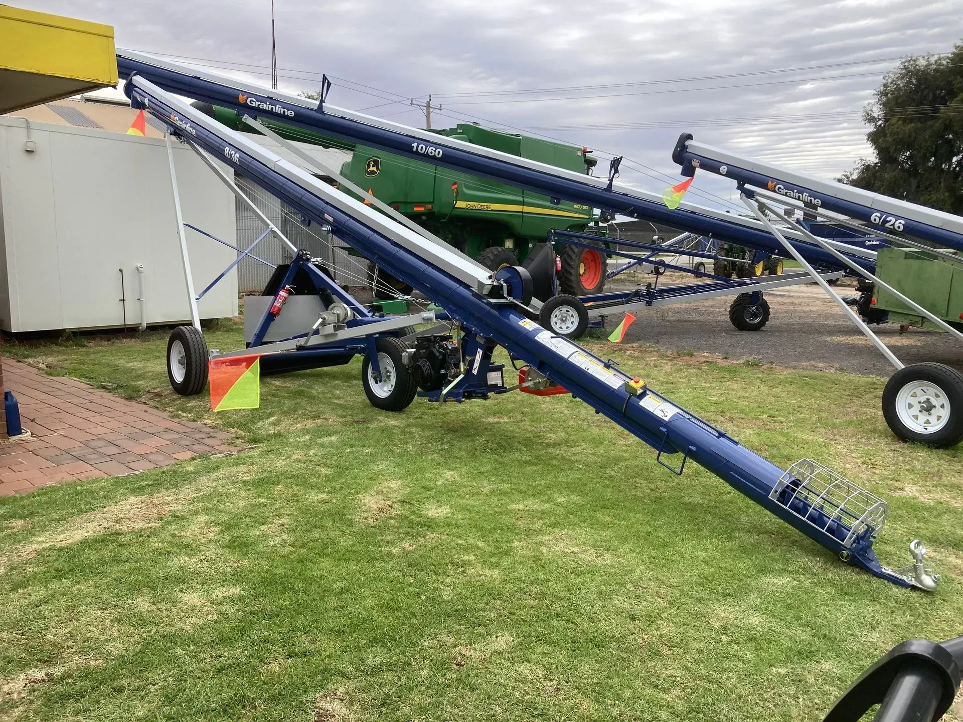 Several blue grain augers on trailers lined up on a grassy lot near a white building