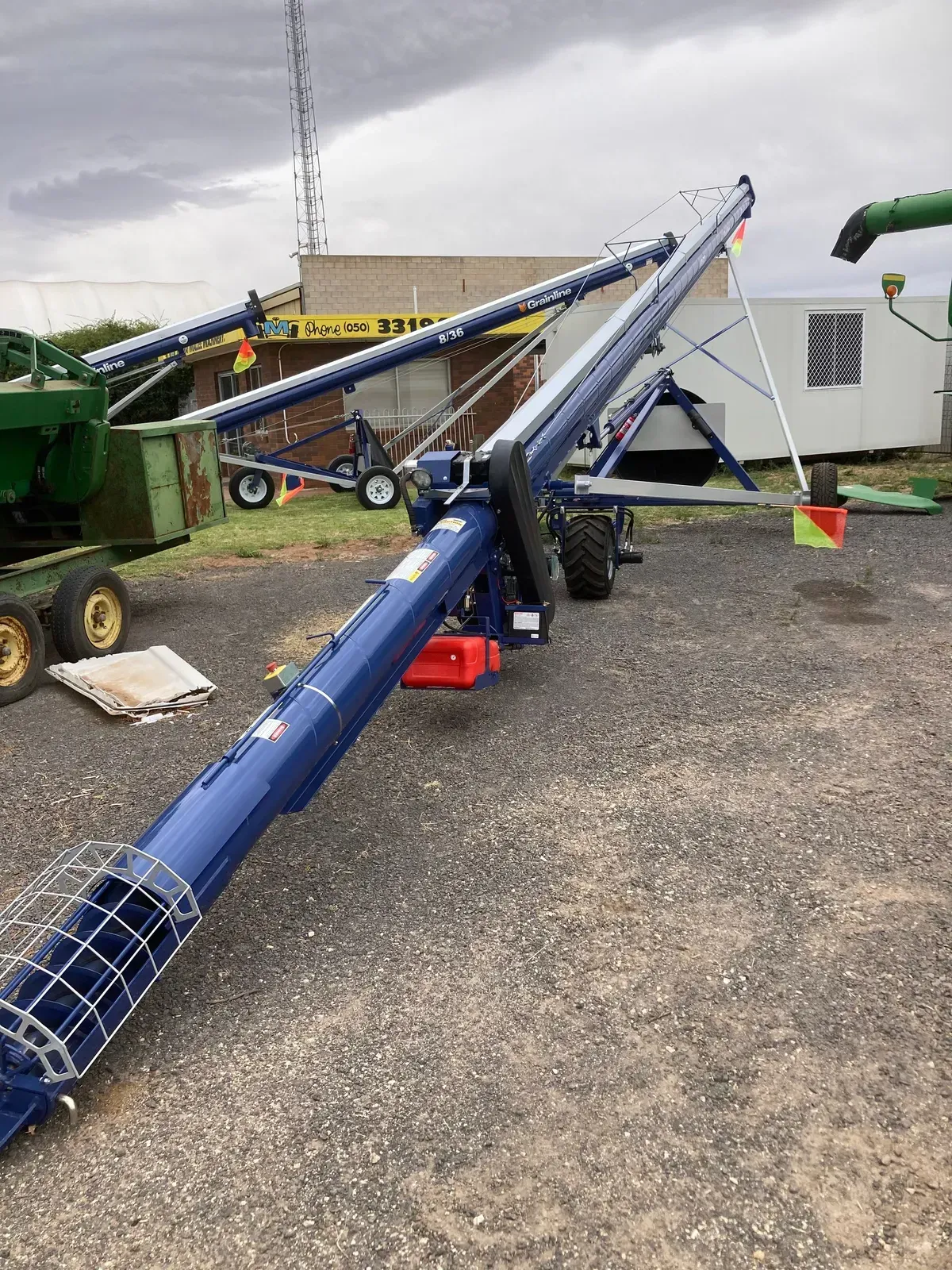 Blue grain auger on gravel beside farm equipment and a white building under cloudy skies