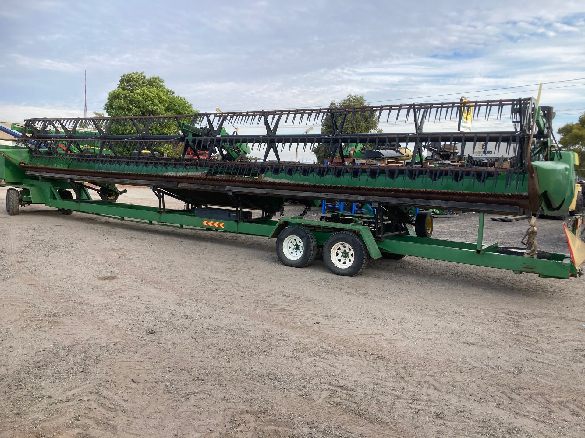 Long green agricultural header on a trailer in a gravel lot