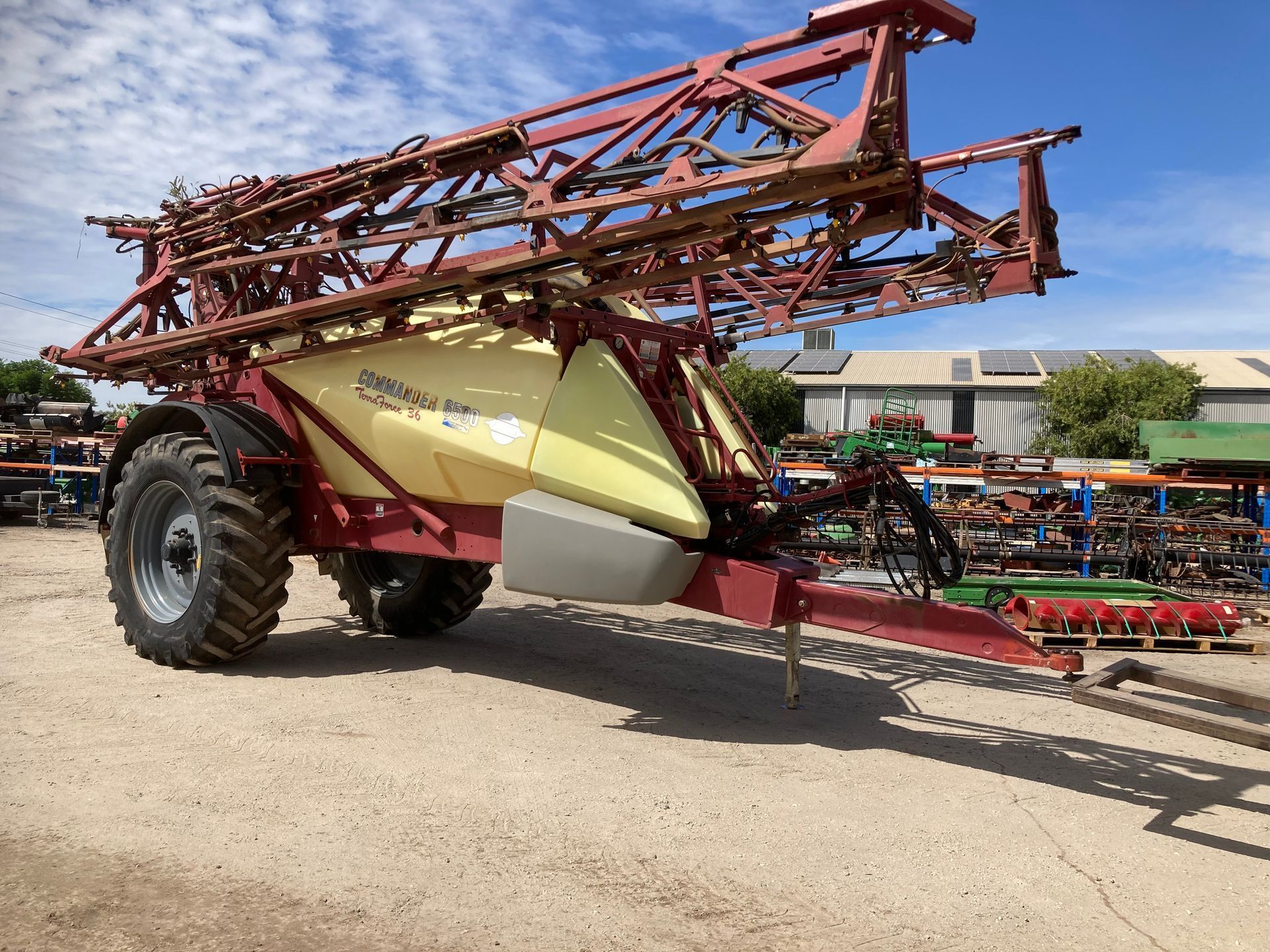 Large agricultural sprayer with folded red booms parked on gravel under a bright sky