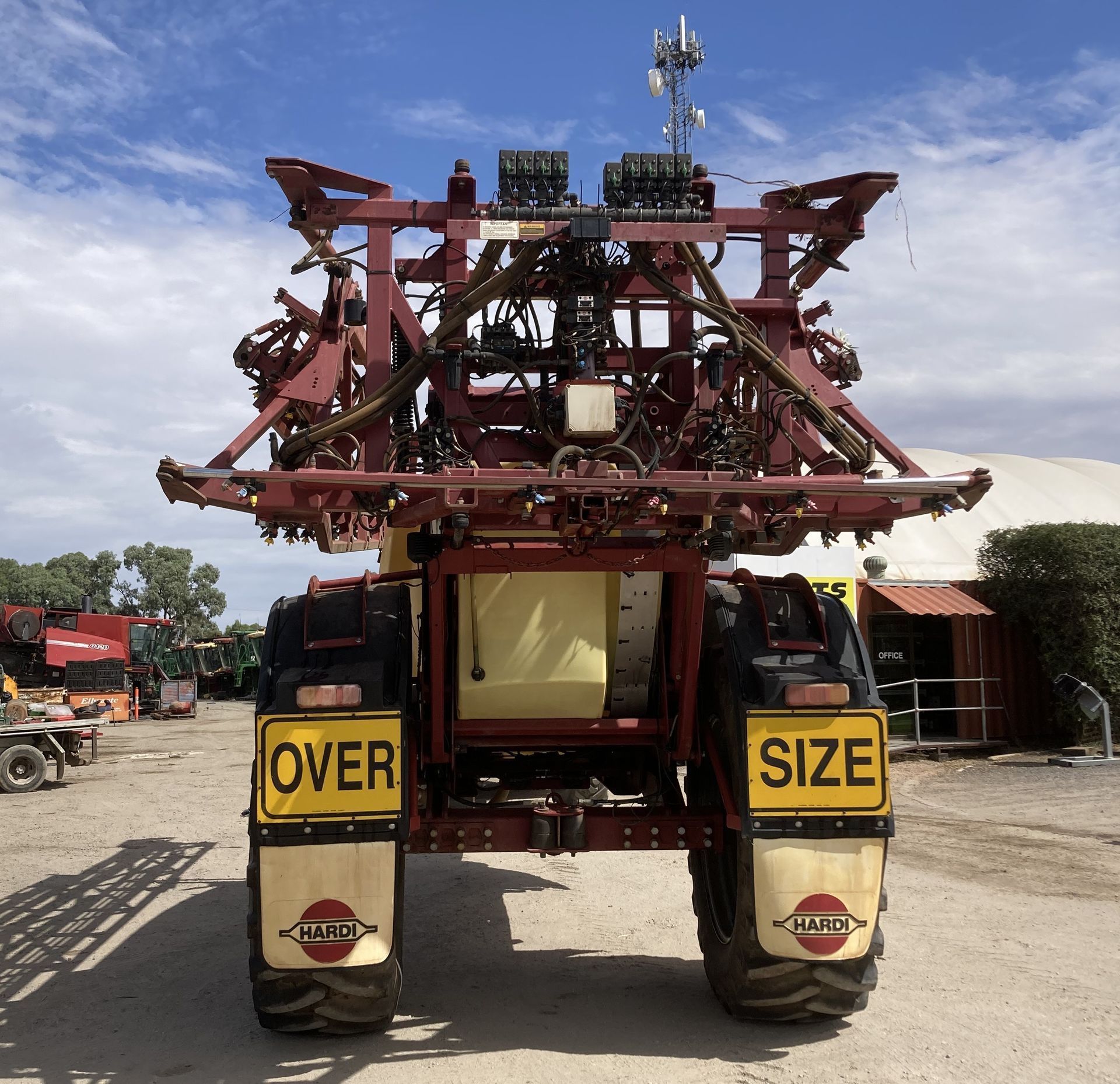 Rear view of a red agricultural machine on a road, with yellow “OVER SIZE” panels and machinery stacked on top