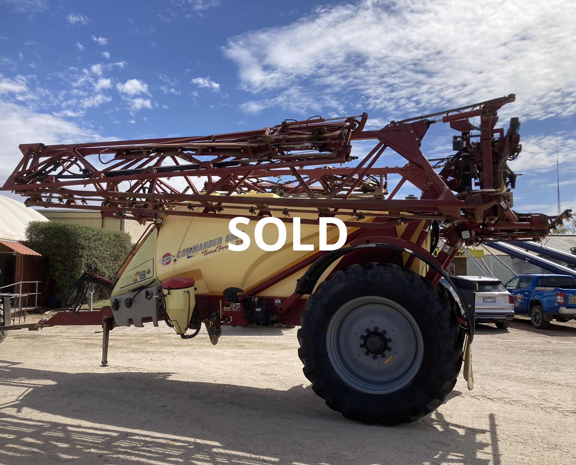 Yellow agricultural sprayer with red boom arms and large tires, parked outdoors with a SOLD sign.