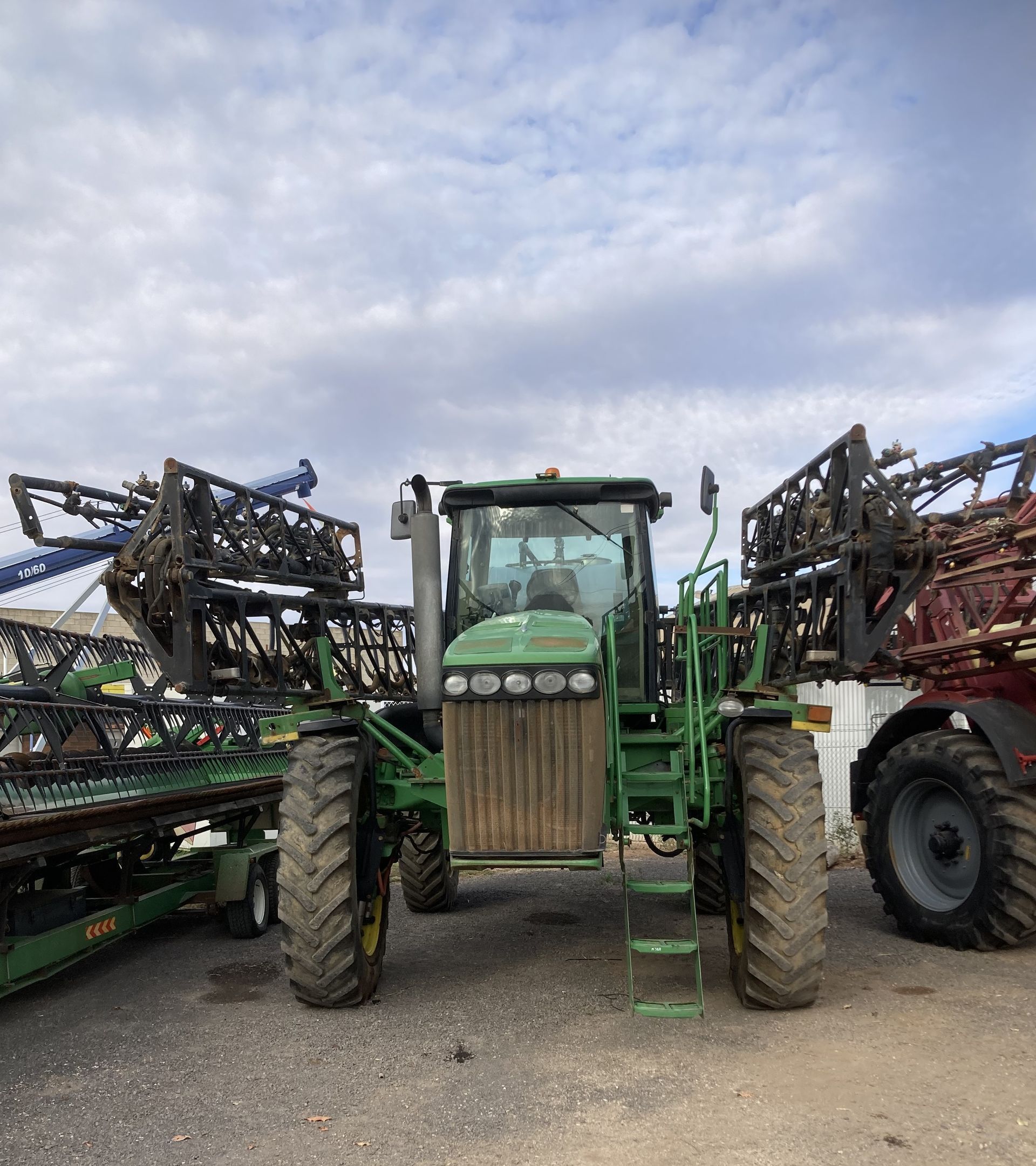 Green tractor between two farm sprayers on a gravel lot under a cloudy sky